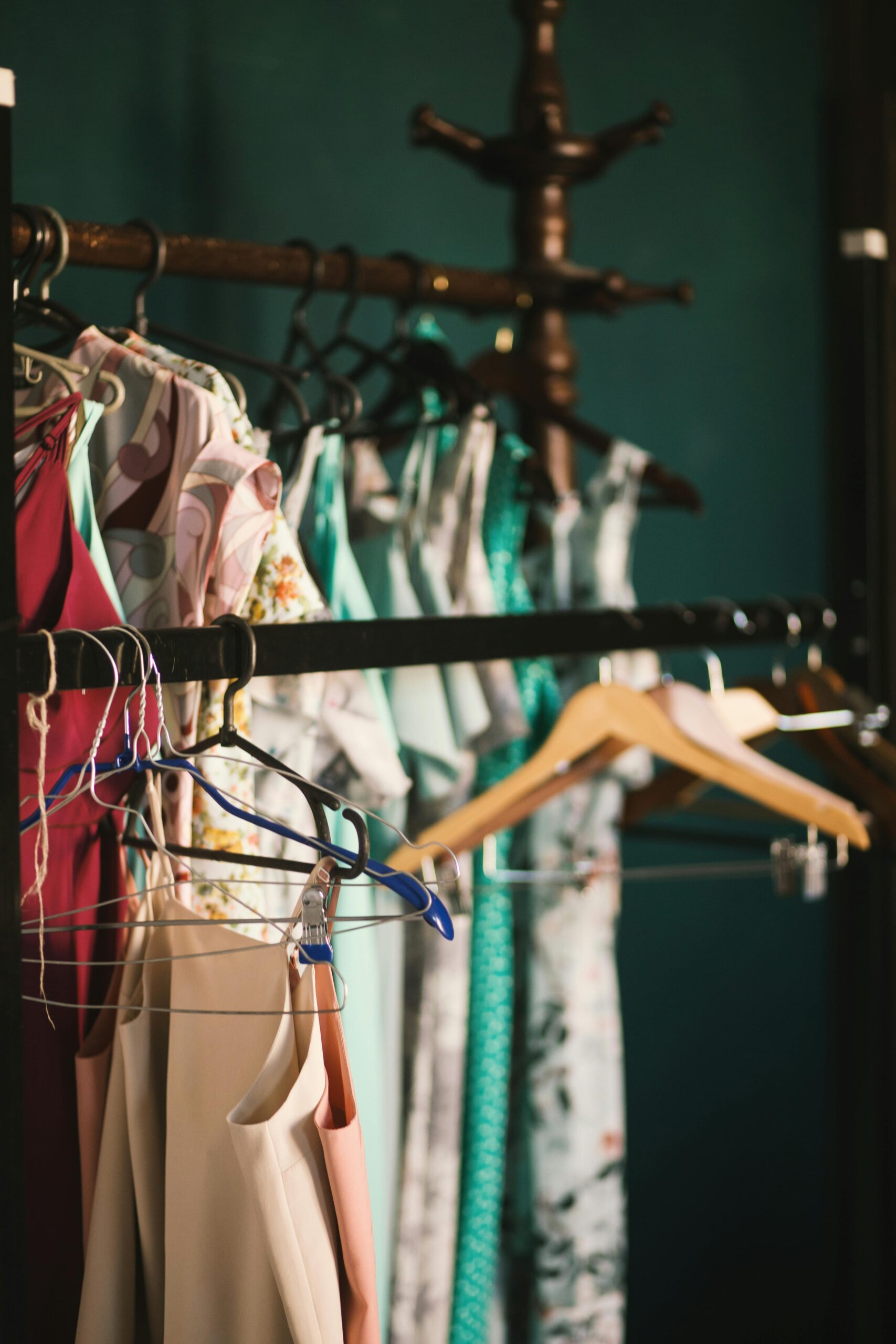 Clothing rack filled with colorful dresses and blouses on various hangers, set against a dark teal background with a vintage wooden coat rack.